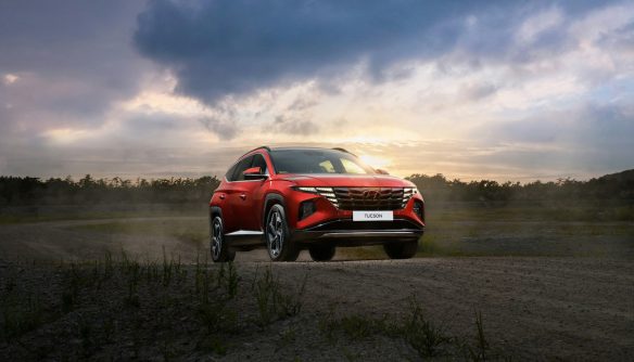 Piros SUV áll egy mezőn, mögötte a lemenő nap megvilágítja a felhőket.