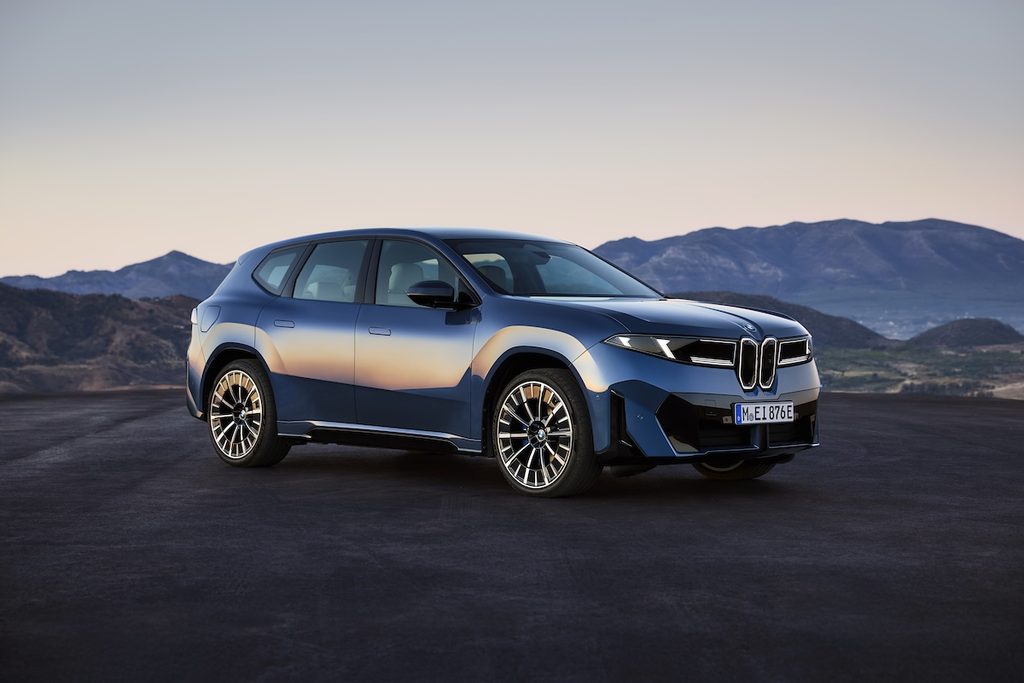 Új sötétkék BMW iX3 50 xDrive, a háttérben hegyek. Az autót megvilágítja a lemenő nap fénye.