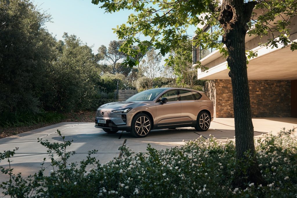 Szürke Volvo EX60 parkol modern ház előtt, növények és fák alatt.