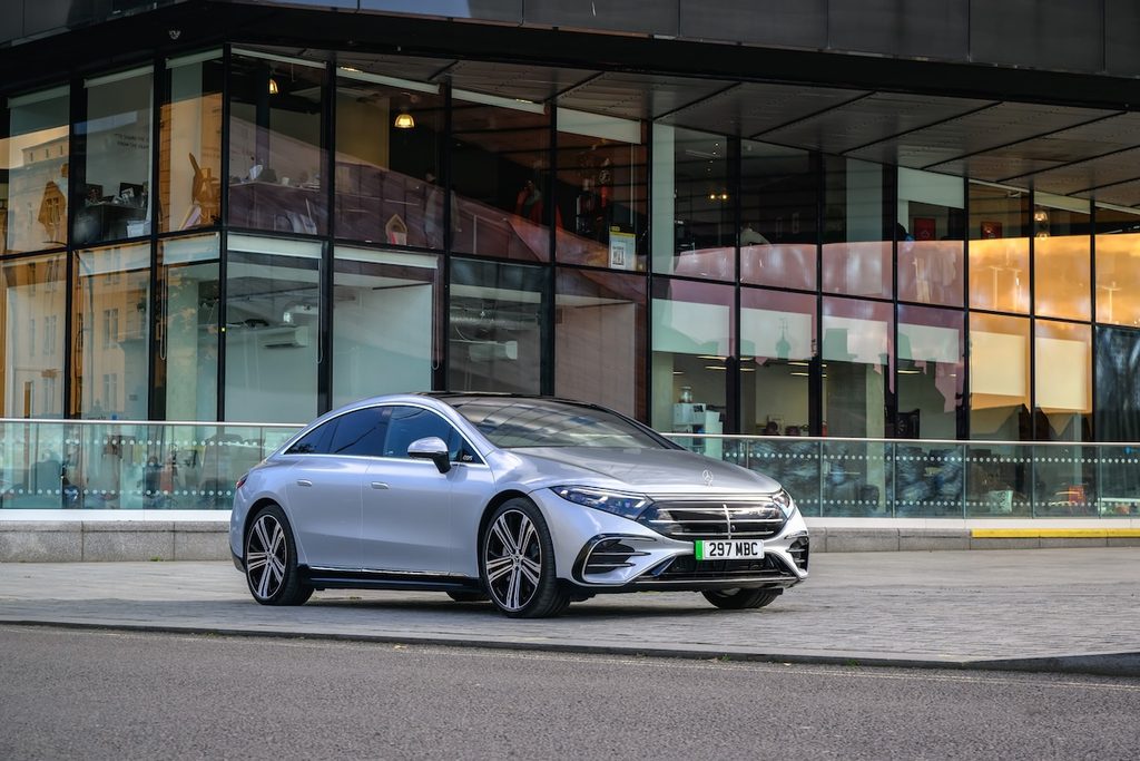 Világosszürke Mercedes-Benz EQS 450+ városi környezetben, modern üveg épület előtt.